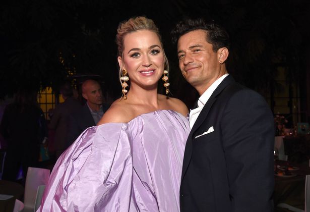 Katy Perry, em um vestido roxo, e Orlando Bloom, em um terno preto, em um tapete vermelho.