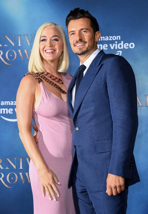 Katy Perry, em um vestido rosa, e Orlando Bloom, em um terno azul, em um tapete vermelho.